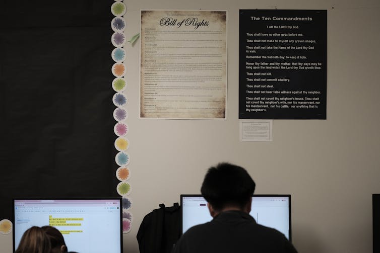 Two students, facing away from the camera, read text on computers positioned up against a white wall.