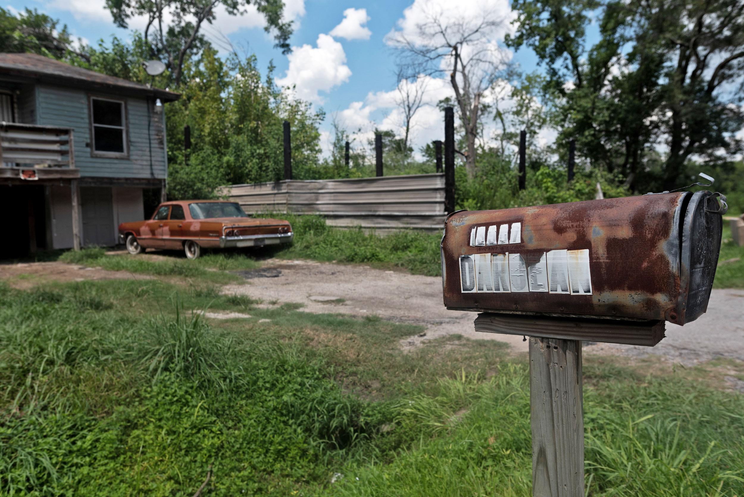 A rusty mailbox with the faded word "DARJEAN" on it stands in front of an aging blue house with red rust-covered car out front