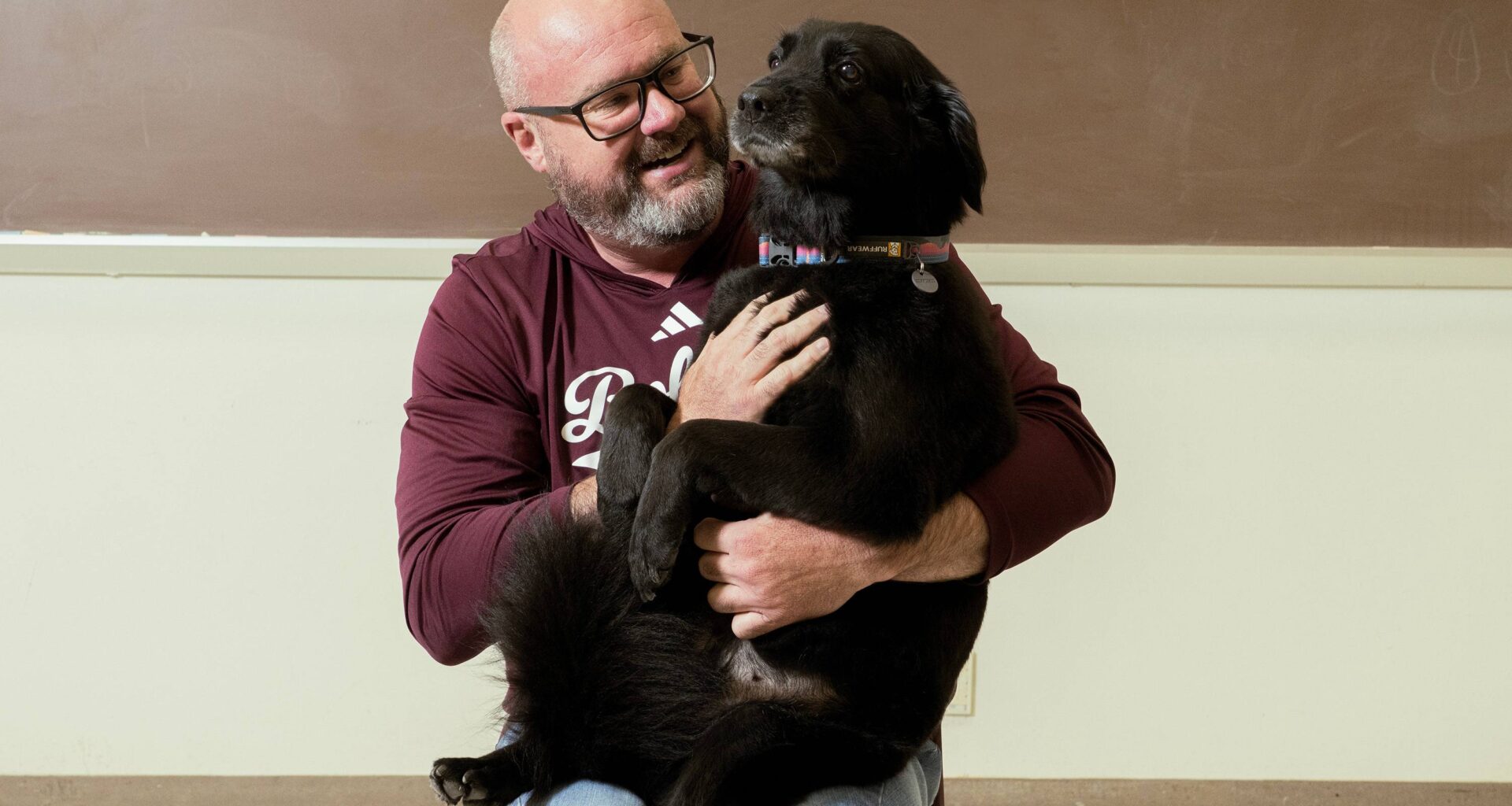 Dale Blasingame with his dog Lucy sitting in his lap like a big baby. Dale is wearing a maroon sweatshirt with Bobcats written on it. Lucy is a medium sized dog with black fur and a little bit of gray on her muzzle.