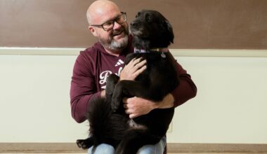 Dale Blasingame with his dog Lucy sitting in his lap like a big baby. Dale is wearing a maroon sweatshirt with Bobcats written on it. Lucy is a medium sized dog with black fur and a little bit of gray on her muzzle.