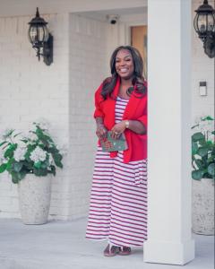 A woman in a red cardigan and striped dress smiles warmly while leaning against a white exterior wall, holding a journal and pen. She wears gold jewelry and her hair is styled in loose waves. White flowers are visible in the background.