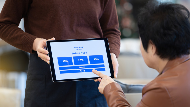 A seated restaurant diner chooses a tip amount on a tablet held by a server in a cafe