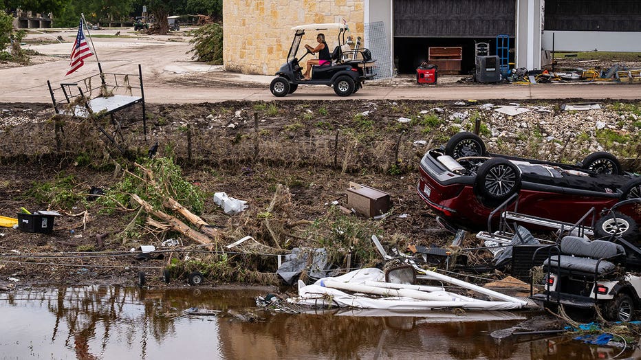 A woman drives past vehicles and golf carts washed out of Buckhorn Lake Resort and onto the shoulder of the Interstate 10 frontage road near Goat Creek Road by flooding, July 17, 2026.