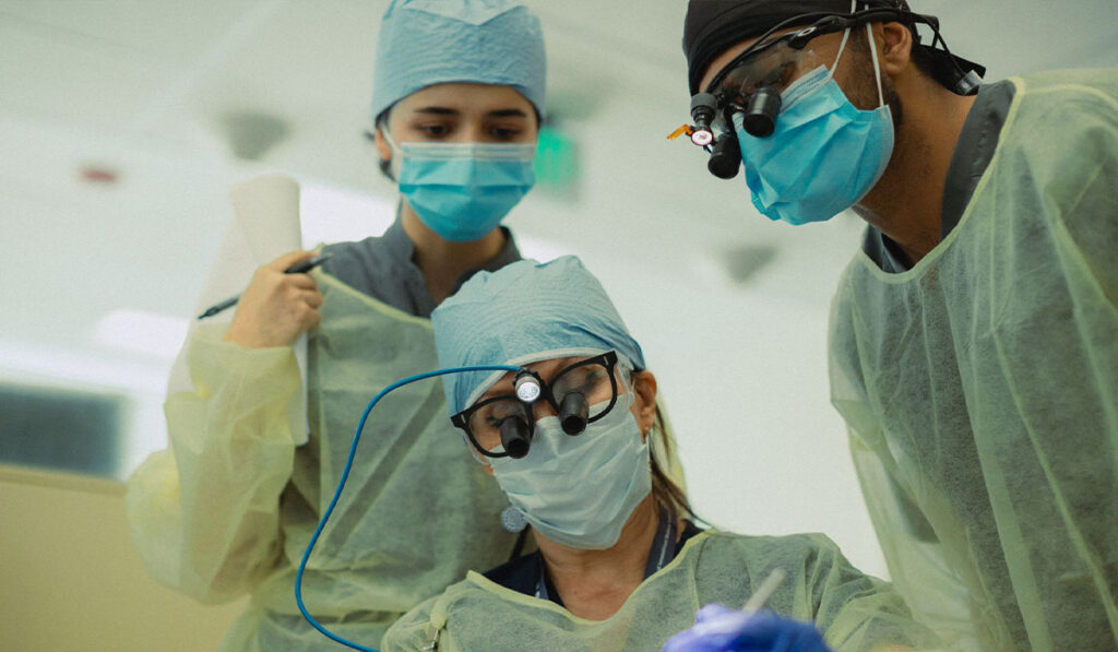 Three people in yellow scrubs and medical caps and masks, lean in to work on someone's teeth.