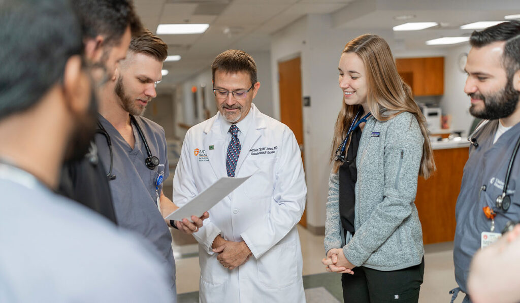 A man in a white medical coat stands in between a group of other people in a hospital setting. A man in grey scrubs reads from a piece of paper as the others listen.