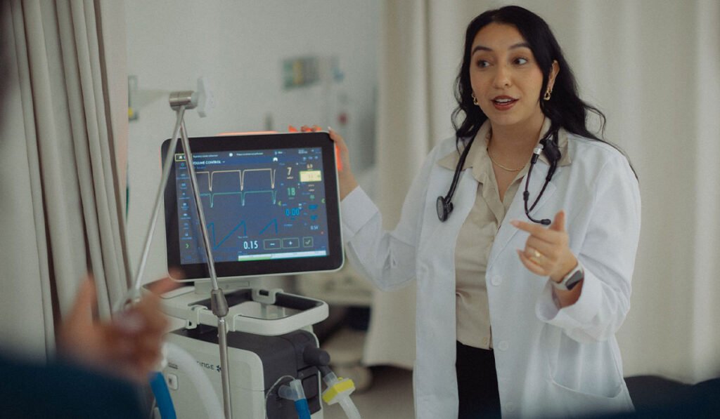 A woman wears a white medical coat with a stethoscope around her neck. She has her hand on a screen with a patient's vital signs.