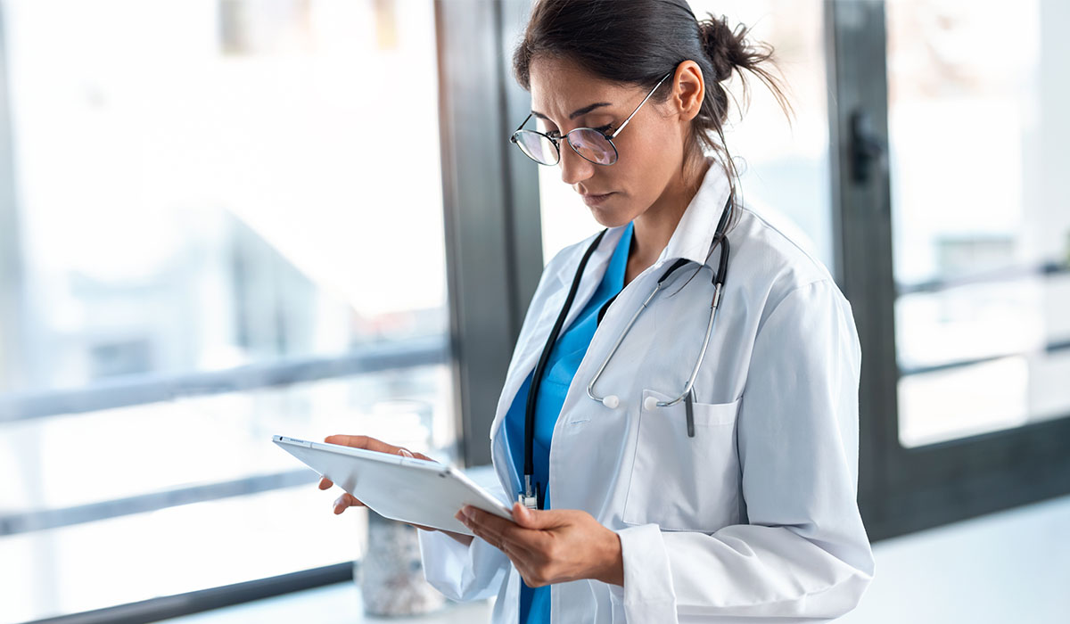 A female doctor examines data on a tablet in a bright hospital hallway.