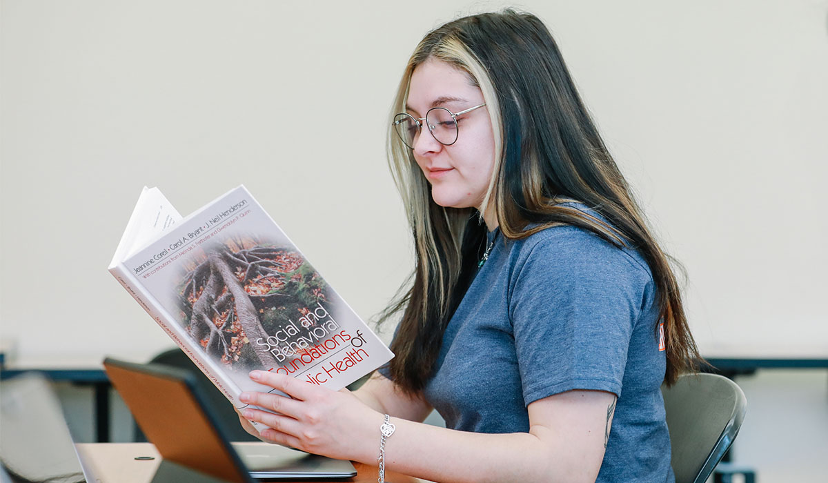 A female student reads a book titled "Social and Behavioral Foundations of Public Health."