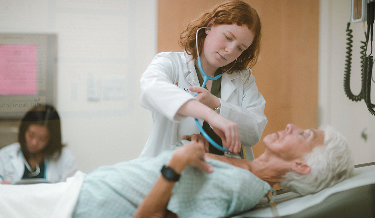 A female medical student holds a stethoscope to the chest of an older female patient lying in a hospital bed.