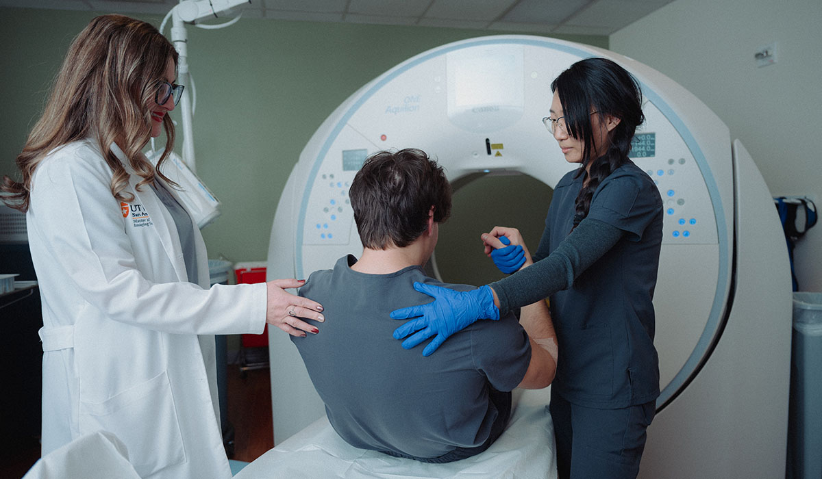 Two women help a male patient rise from a laying position after an MRI.