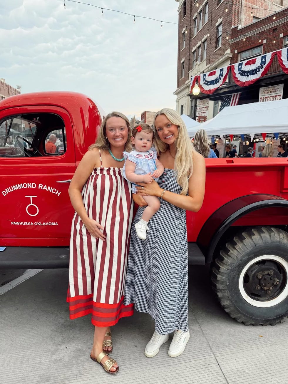 Two women and a baby stand together in front of a vintage red truck at a festive outdoor event. Two women and a baby stand together in front of a vintage red truck at a festive outdoor event.