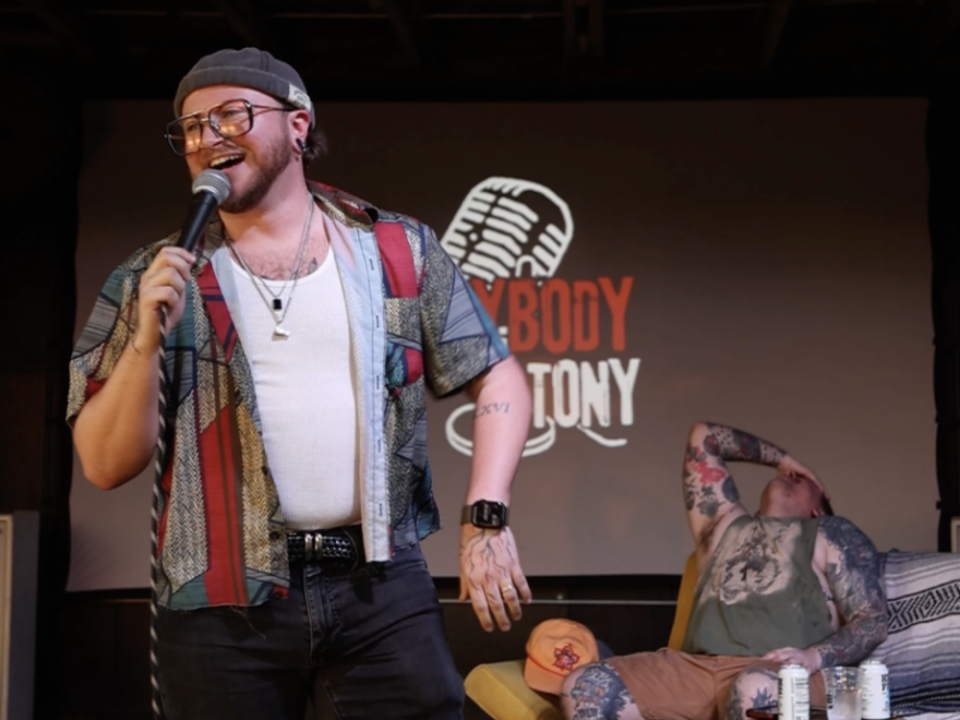 Anybody But Tony co-host Everett Bragg does stand-up while Erik Holden laughs on a couch behind him.