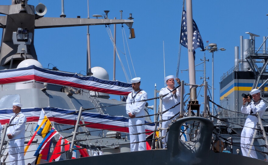 Photo of the bow of the USS Fort Worth where sailors in dress white uniforms stand at ease while one strikes the colors — removing the commissioning flag of the ship.