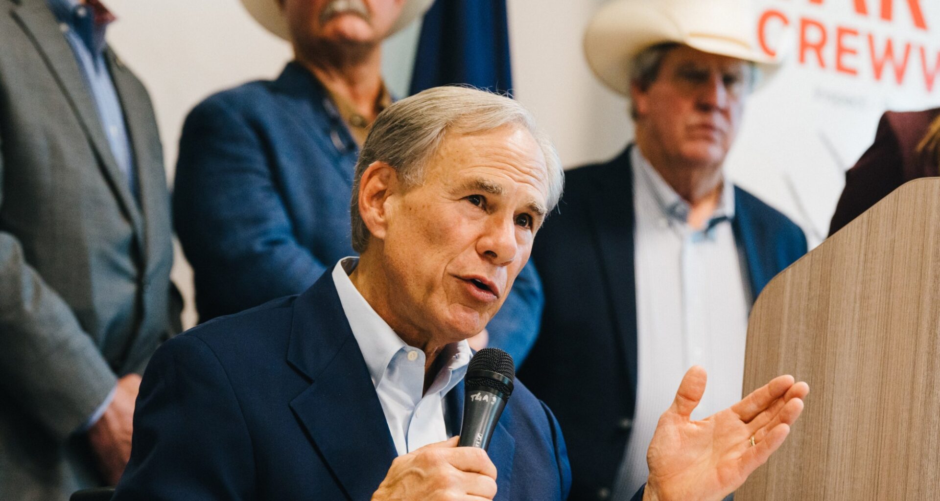 Texas Gov. Greg Abbott speaks into a microphone with his left hand raised in a gesture, addressing an audience. He is dressed in a navy suit jacket over a light-blue collared shirt.