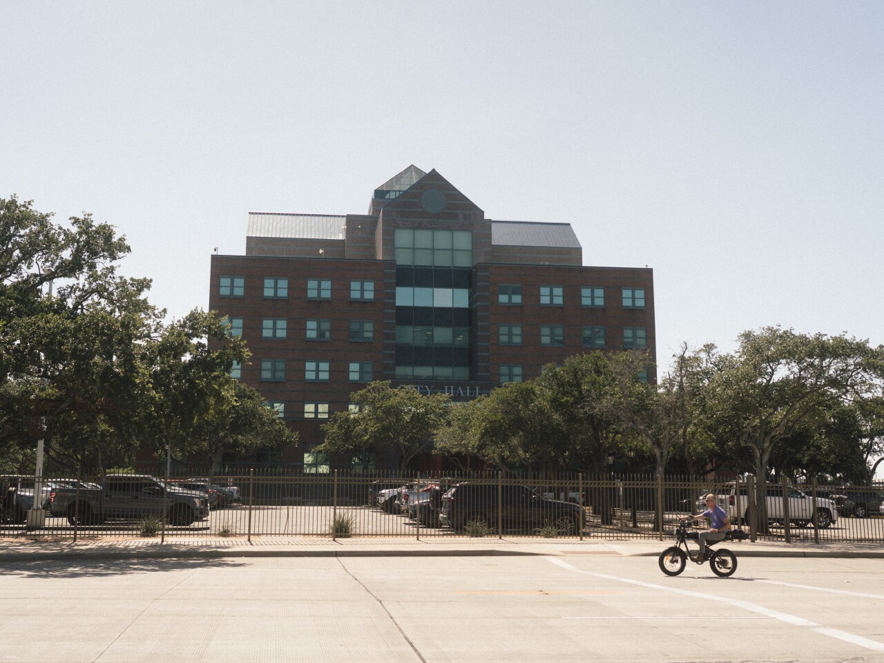 A wide shot shows a multistory brick building labeled "City Hall" behind a fenced-in parking lot. In the foreground, a person rides a black bicycle across the wide, paved street under a bright clear sky.