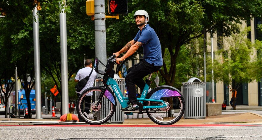 A man rides a Bikeshare bike along a downtown Austin street. The bike emblazoned with the word "CapMetro." 