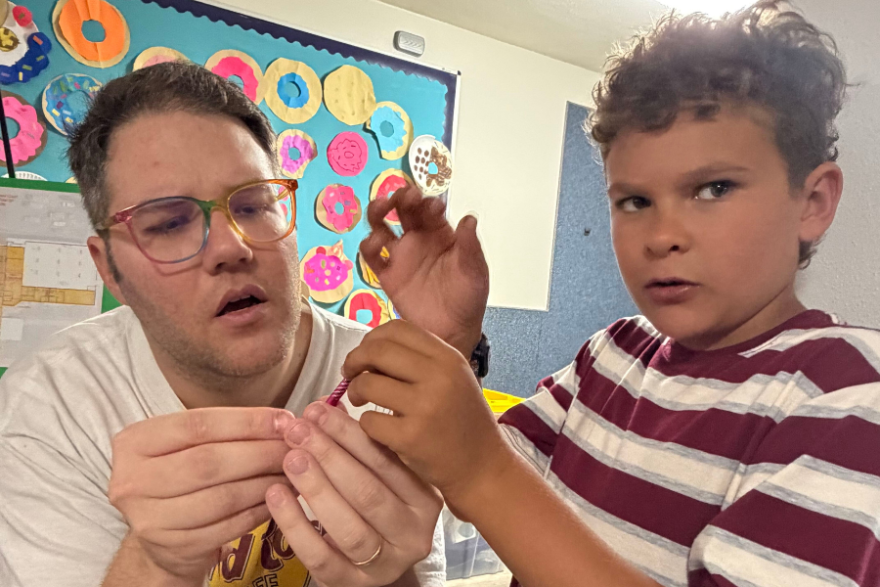 A man in a Hard Rock Cafe t-shirt and colorful glasses helps a young boy in a striped shirt with a colored pencil. They are in a classroom with colorful paper donuts hanging on the wall behind them.