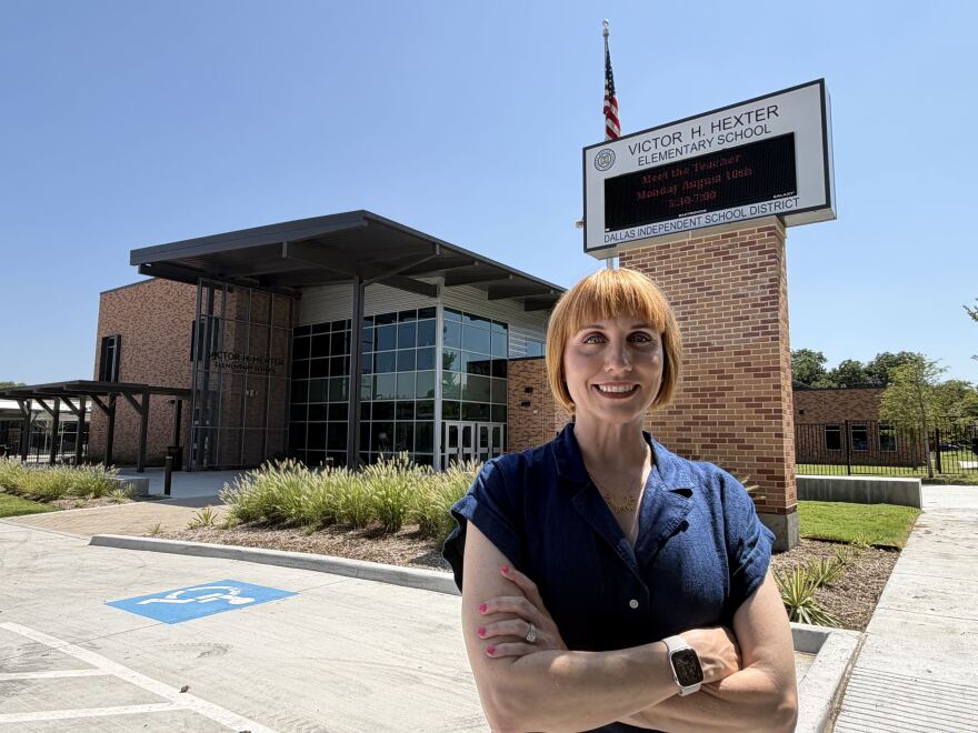 A white woman with a short red bob wearing a blue shirt stands outside a brick and glass school building. On a sign behind her it says "Victor H. Hexter Elementary School."