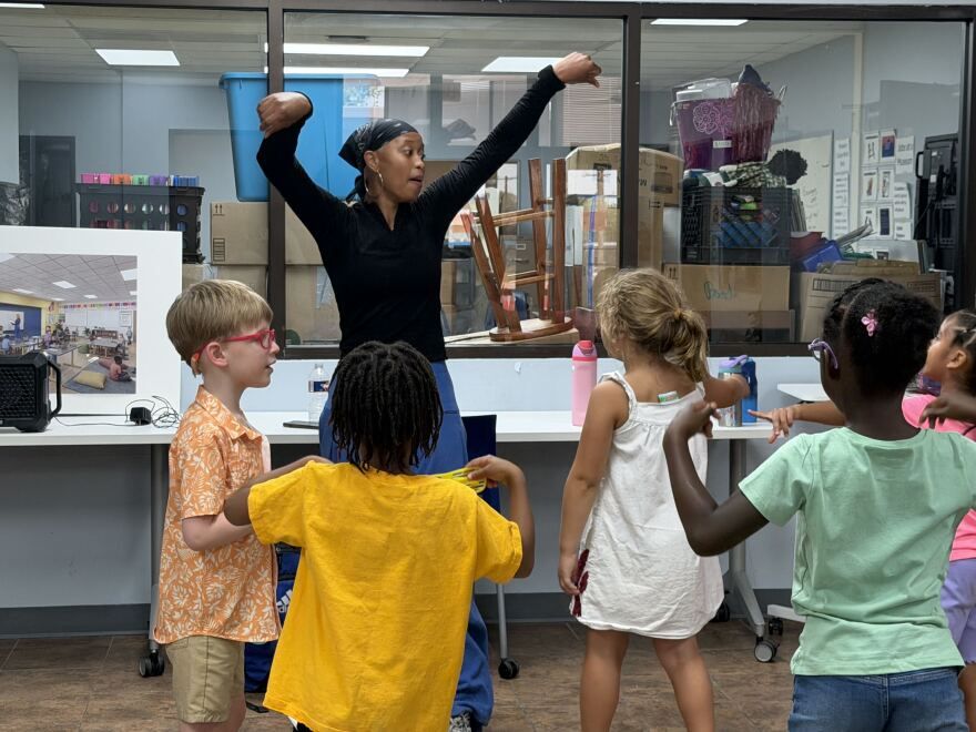 A Black woman in a black top and blue pants leads a group of young kids in a dance class. 