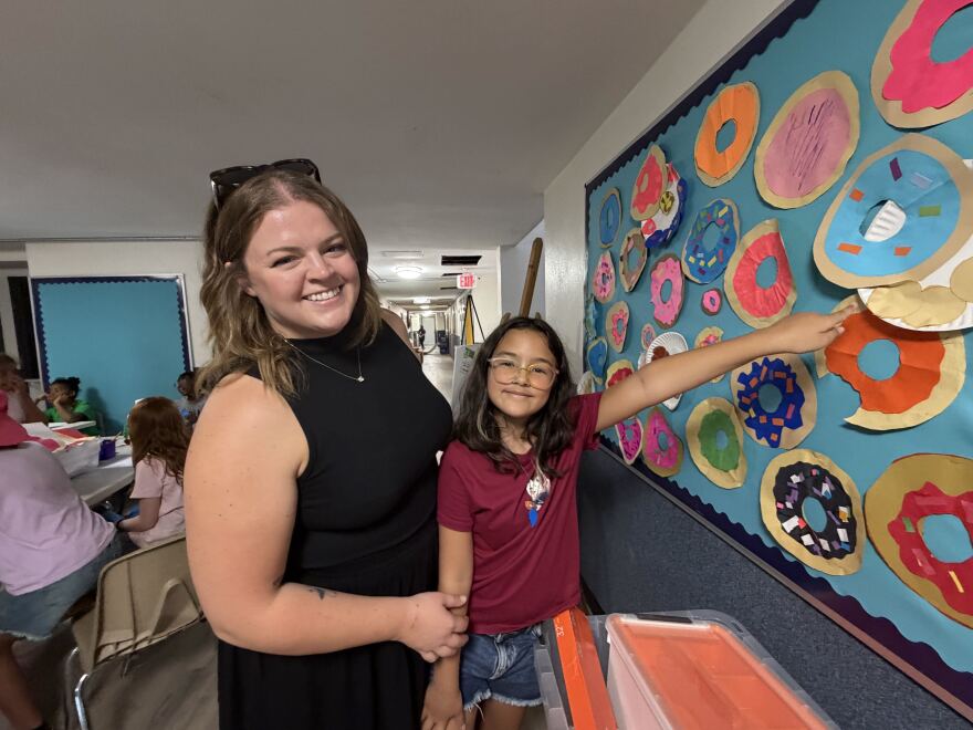 A woman in a black top stands next to her 9-year-old daughter, in a red top and glasses, as she points to a paper donut hanging on the wall of a classroom. They are both smiling at the camera. 