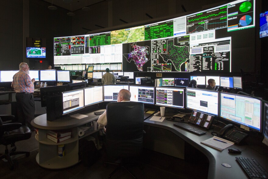 Two men in the foreground sit at desks with more than a dozen monitors on them while a wall in the background is covered with more large screens with maps of Texas and various data streams on electricity across the state.