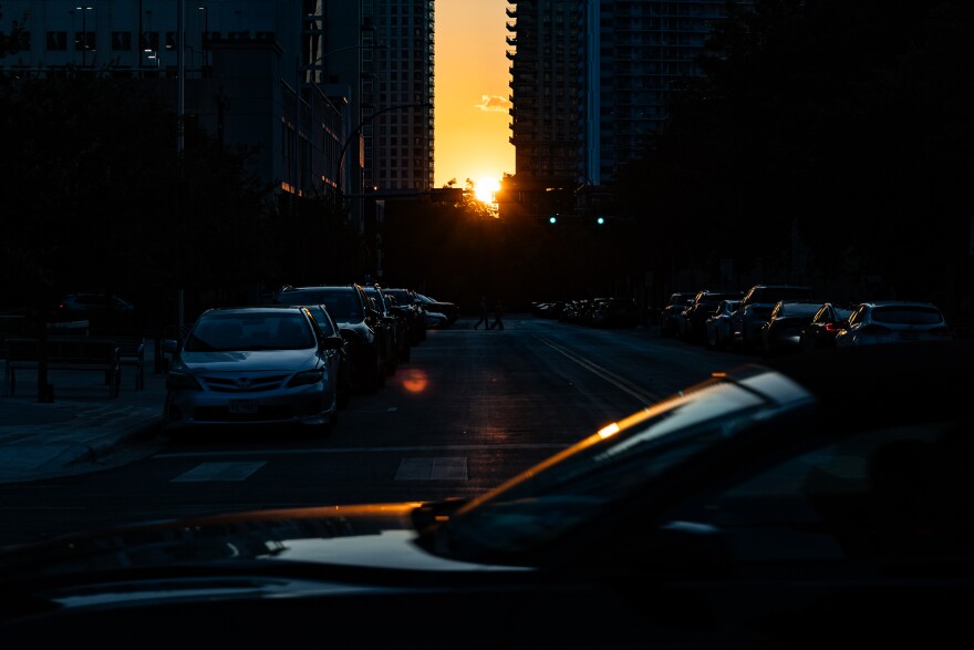 A city street at dusk, with the sun setting between tall buildings, casting a warm glow over parked cars and trees.