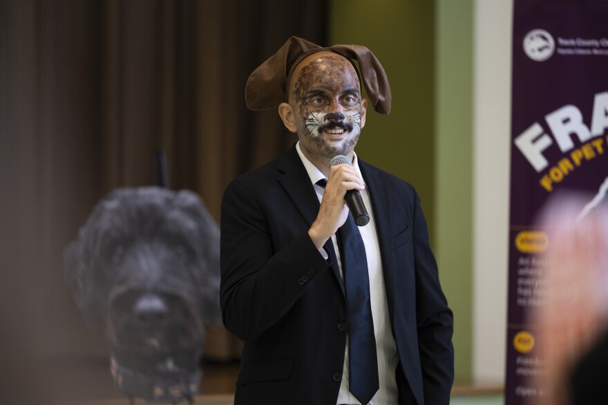 A man wearing face paint that resembles a dog speaks into a microphone with a large cutout photo of a dog behind him. 