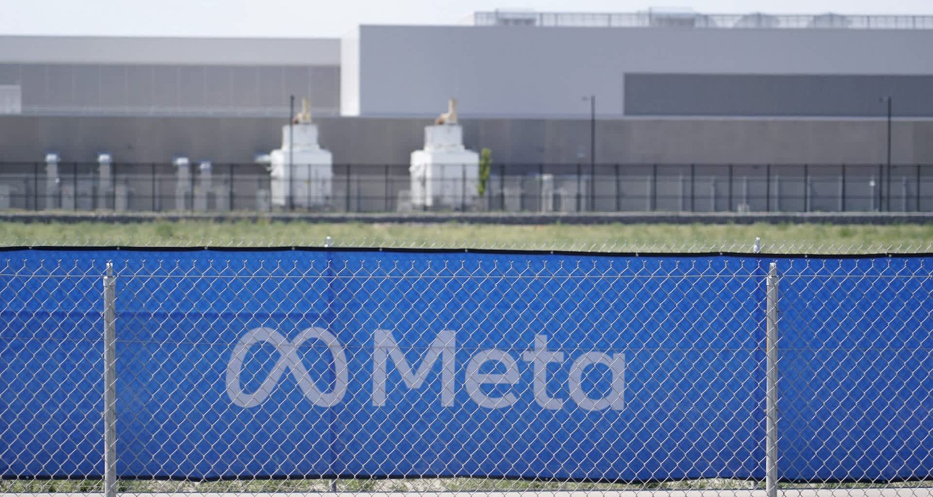 A blue fence with the Meta logo and a background of industrial buildings and green fields.