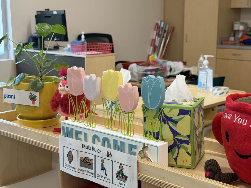A teacher's desk in a pre-kindergarten classroom. It is covered in a tissue box, plus heart, wooden flowers over the word "welcome" and a plant in a yellow pot.