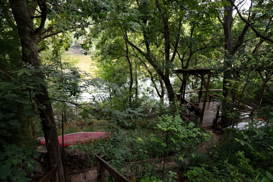 From a raised vantage point a pathway leads away to a river bank, surrounded by dense green foliage and trees. In the corner of the photo is a red watercraft resting in the grass.
