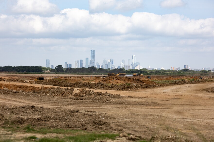 Construction vehicles drive across the churned earth of a cleared construction site as the cityscape of downtown Austin sits on the horizon.