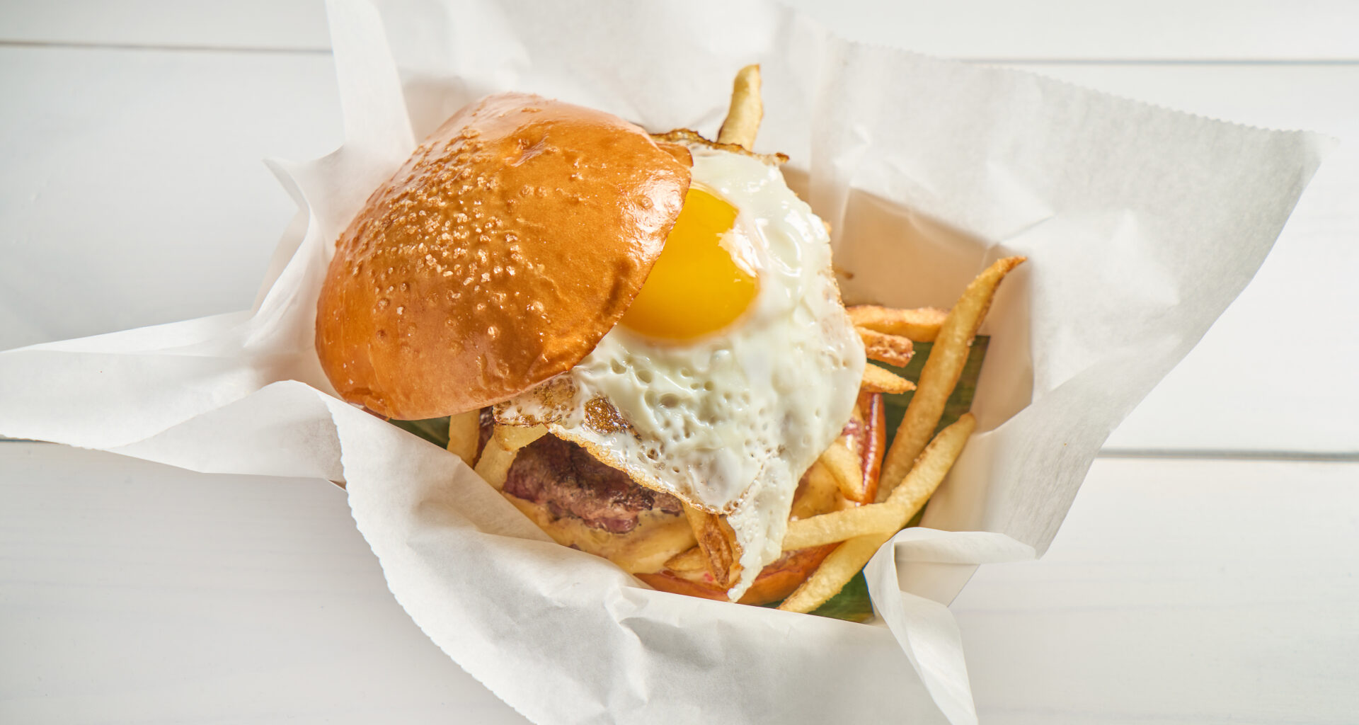 Frita Batidos chorizo burger topped with a fried egg and crispy shoestring fries on a toasted bun, served in a paper-lined basket.