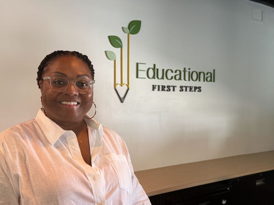 A woman in a white button-down shirt and glasses with hoop earrings stands in front of a wall with the words "Educational First Steps" on it. Its logo is a yellow pencil with leaves on the top.