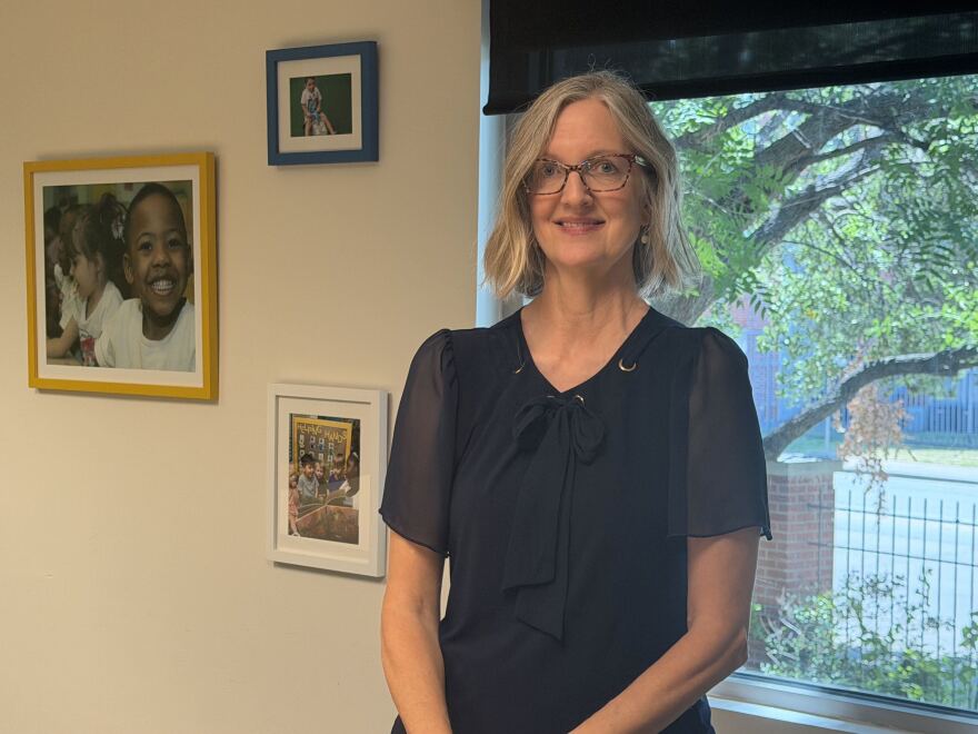 A woman in a dark blouse stands in an office in front of a window. She has shoulder-length blonde hair and wears glasses.