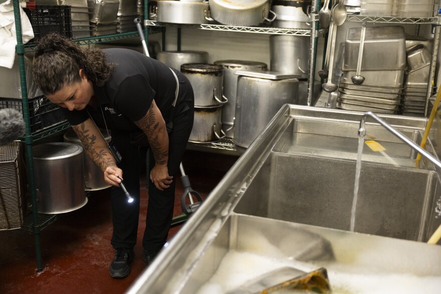 A woman in black uses a flashlight to look under sinks in the back of a restaurant kitchen.