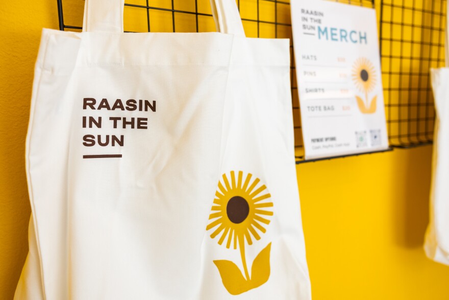 A bag that reads Raasin in the Sun with a sunflower on it hangs from a wire shelf against a yellow background.