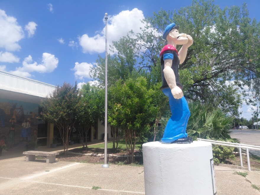 A Popeye statue stands in front of a building with bright mural. Sidewalk, trees and rails are in the background.