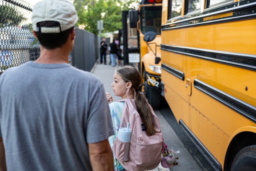 A person and a child with a pink backpack stand on a sidewalk beside a yellow Austin Independent School District school bus.
