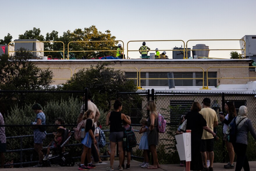 People line up along a fenced school as workers in safety vests stand on a rooftop above.