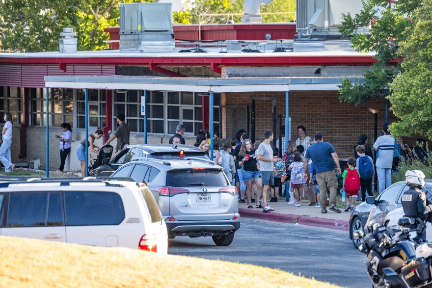 Parents and children mill around the outside of of a school building during student drop off in the morning.
