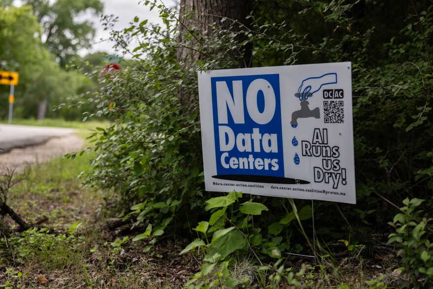 A white and blue sign that reads "No Data Centers. AI Runs Us Dry" with an image of a water faucet sits on a roadside amid green foliage.