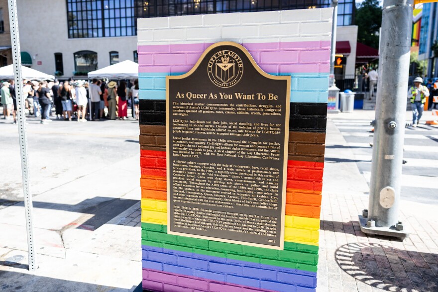 A metallic City of Austin historical marker titled "As Queer As You Want To Be" sits on a brick pillar painted in a rainbow of colors.