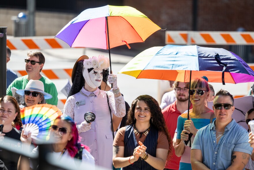 A crowd of smiling people, one dressed in drag, hold rainbow colored fans and umbrellas while listening to a speaker out of frame.
