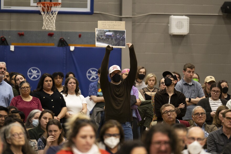 A man in a black mask holds up a sign with photograph of a vehicle surrounded by law enforcement. The sign reads "APD After Running Your Neighbor Off The Road."