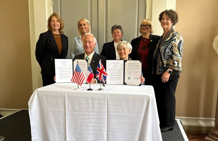 A group of people pose for a photo celebrating the signing of a sister city agreement. A table with a white tablecloth is at the center with small tabletop flags representing the United States, Texas and Greater Manchester.