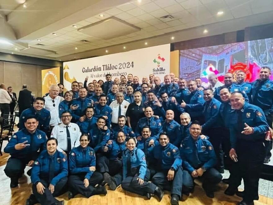 A large group of firefighters wearing blue uniforms pose for a photo. The firefighters are from Saltillo, Mexico.