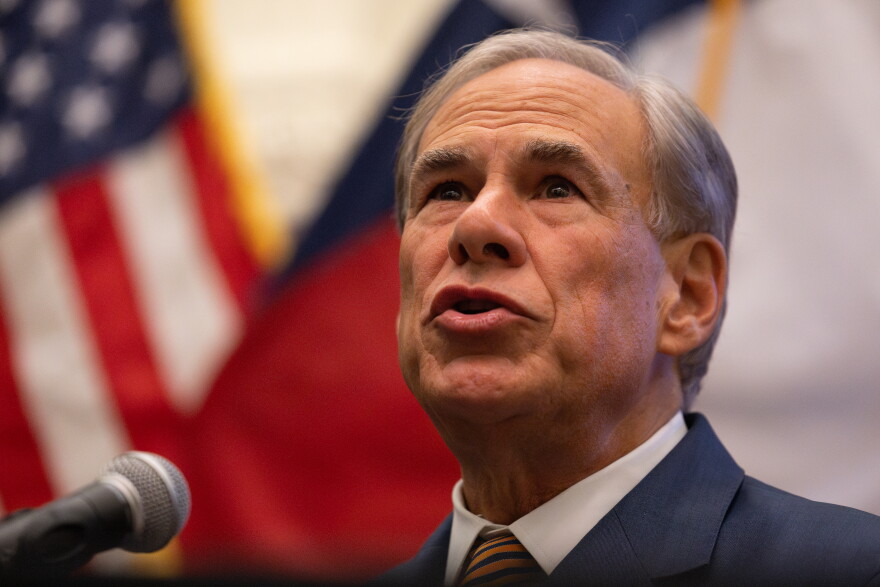 A close up of a man in a suit speaking into a microphone in front of Texas and United States flags.