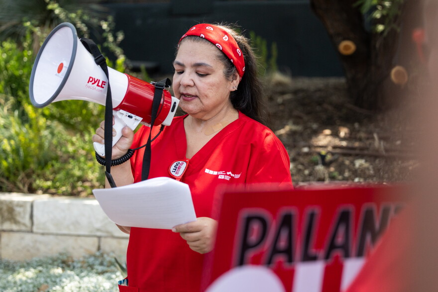 A person in a red outfit and headband speaks into a megaphone while holding papers, with a partially visible red sign in the foreground.