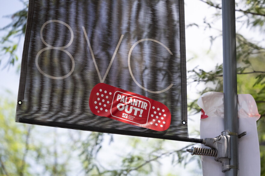 A black banner with "8VC" and a red bandage sticker reading "Palantir Out!" is attached to a pole outdoors.