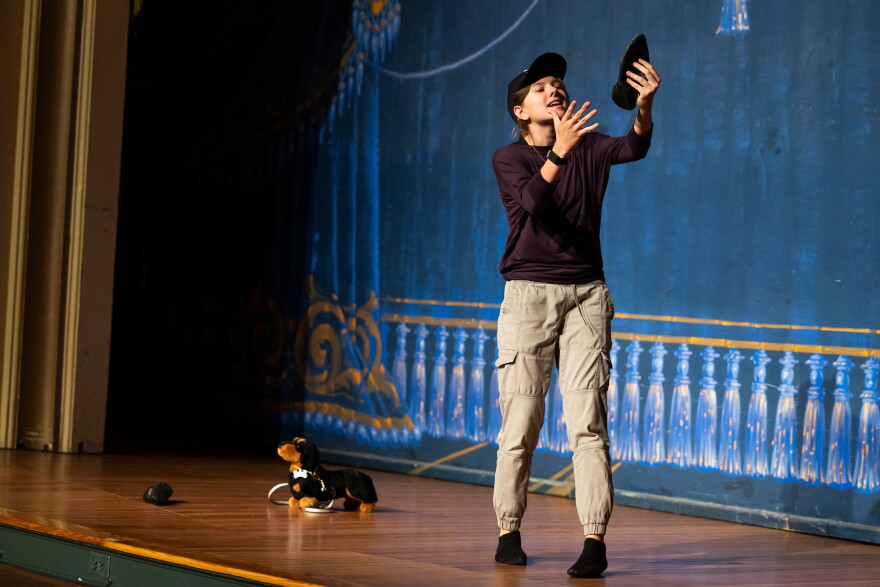 Skylar Ewing holds up a shoe on stage as she performs a scene from Two Gentlemen of Verona. A stuffed dog is on the floor beside her.